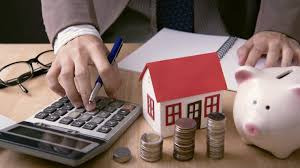 Kalkulator, model domu z czerwonym dachem, monety ułożone w stosy, świnka skarbonka, okulary i notatnik na biurku. Osoba w garniturze używa kalkulatora.