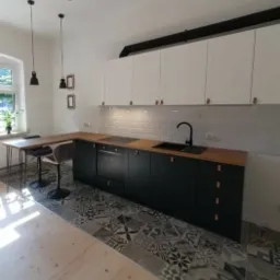 Nowoczesna kuchnia z czarnymi szafkami dolnymi, drewnianym blatem i białymi szafkami górnymi, wzorzysta podłoga, oświetlenie punktowe i wiszące lampy.