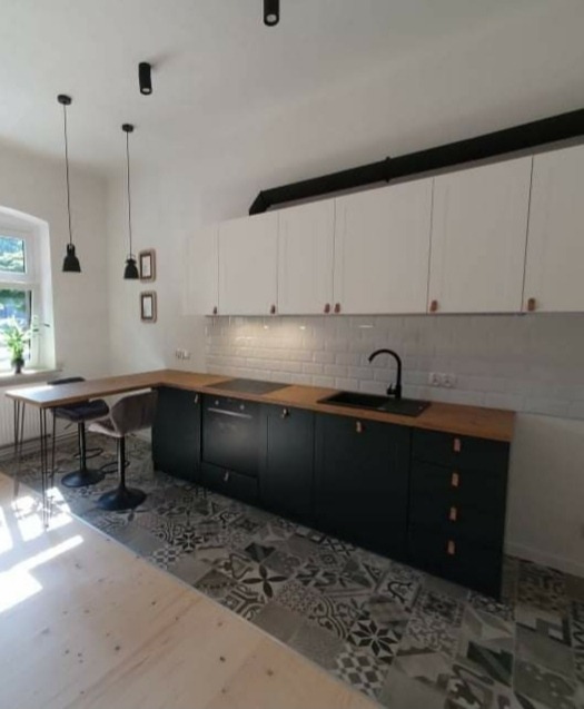 Nowoczesna kuchnia z czarnymi szafkami dolnymi, drewnianym blatem i białymi szafkami górnymi, wzorzysta podłoga, oświetlenie punktowe.