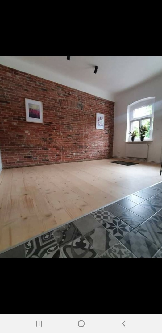 Surowe deski podłogowe przechodzące w płytki ceramiczne z wzorami, ściana z czerwonej cegły z dwoma obrazami, okno z roślinami, czarne lampy sufitowe.