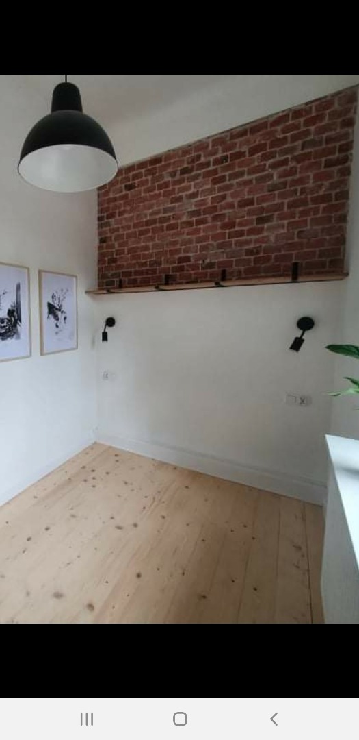 Pokój z fragmentem ściany z czerwonej cegły, drewniana podłoga, czarna lampa wisząca i dwa kinkiety, półka z czarnymi wspornikami, minimalistyczne grafiki na ścianie.