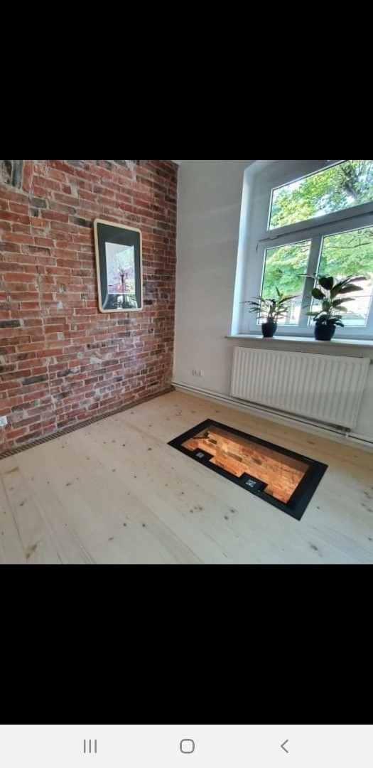 Pokój z odsłoniętą ceglaną ścianą, jasną drewnianą podłogą i oknem z roślinami; w podłodze okno z widokiem na ceglany piwniczny sufit.