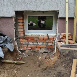 Wstawianie okna w mur z czerwonej cegły, widoczny fragment elewacji, poziomica i wykop ziemny.