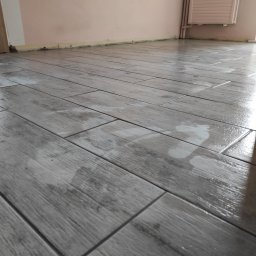 Super Mario Usługi Remontowo-Budowlane - Ułożone szare płytki podłogowe imitujące drewno, świeżo po umyciu, z widocznymi smugami wody, na tle ściany w kolorze brudnego różu i grzejnika.