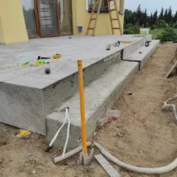 Układanie płytek gresowych na schodach zewnętrznych tarasu, widoczne narzędzia glazurnicze i elementy mocujące, drabina oparta o elewację budynku.