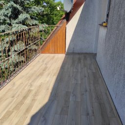 Super Mario Usługi Remontowo-Budowlane - Balkon z nową podłogą z płytek imitujących drewno, żeliwna balustrada z ozdobnym wzorem, widok na drzewa i antenę satelitarną.