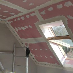 Super Mario Usługi Remontowo-Budowlane - Poddasze w trakcie ocieplania, widoczne płyty gipsowo-kartonowe z zaszpachlowanymi łączeniami i otwartym oknem dachowym, drabina malarska w rogu pomieszczenia.