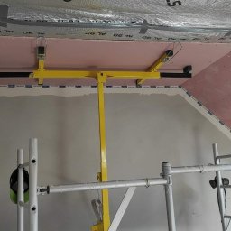 Super Mario Usługi Remontowo-Budowlane - Montaż płyt gipsowo-kartonowych na poddaszu z użyciem żółtego podnośnika, widoczne rusztowanie i izolacja termiczna.