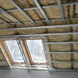 Poddasze w trakcie ocieplania z widoczną izolacją z wełny mineralnej, metalową konstrukcją pod płyty gipsowo-kartonowe i dwoma oknami dachowymi.