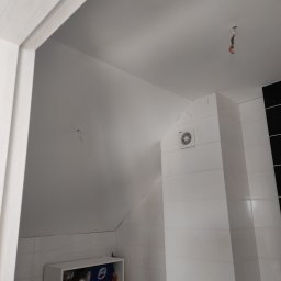 Super Mario Usługi Remontowo-Budowlane - Łazienka po remoncie: białe płytki na ścianach i zabudowie, wentylator w narożniku, pod skosem sufitu półka z artykułami chemicznymi, wystające kable z sufitu.