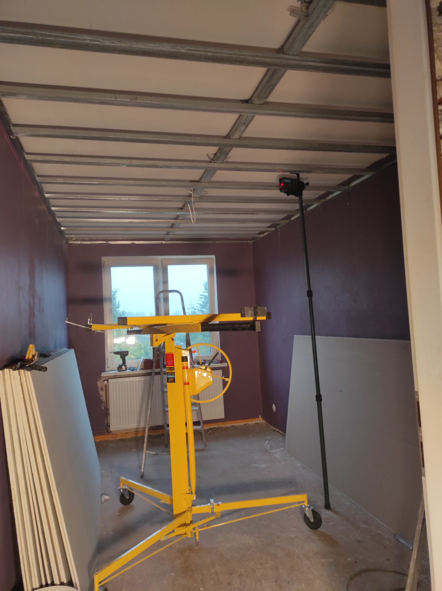 Wnętrze pokoju z purpurowymi ścianami w trakcie montażu sufitu podwieszanego z metalową konstrukcją i płytami gipsowo-kartonowymi, widoczny żółty podnośnik do płyt oraz laserowy niwelator na statywie.