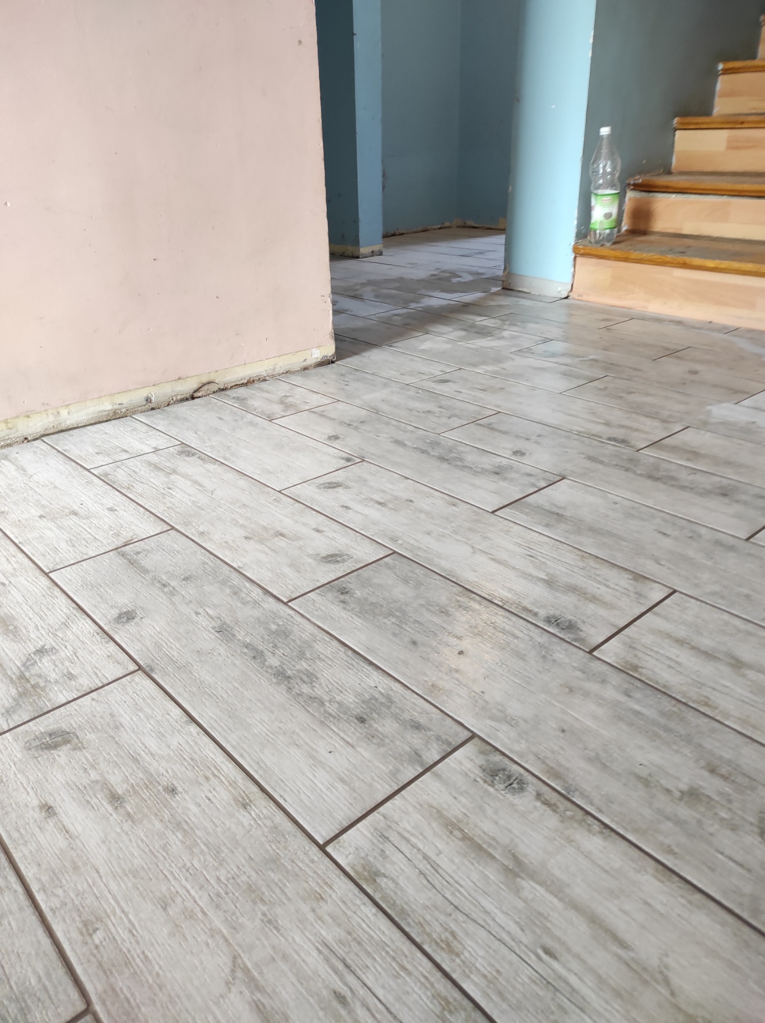 Ułożona podłoga z płytek imitujących drewno, widoczne schody i ściana w trakcie remontu, butelka wody przy schodach.
