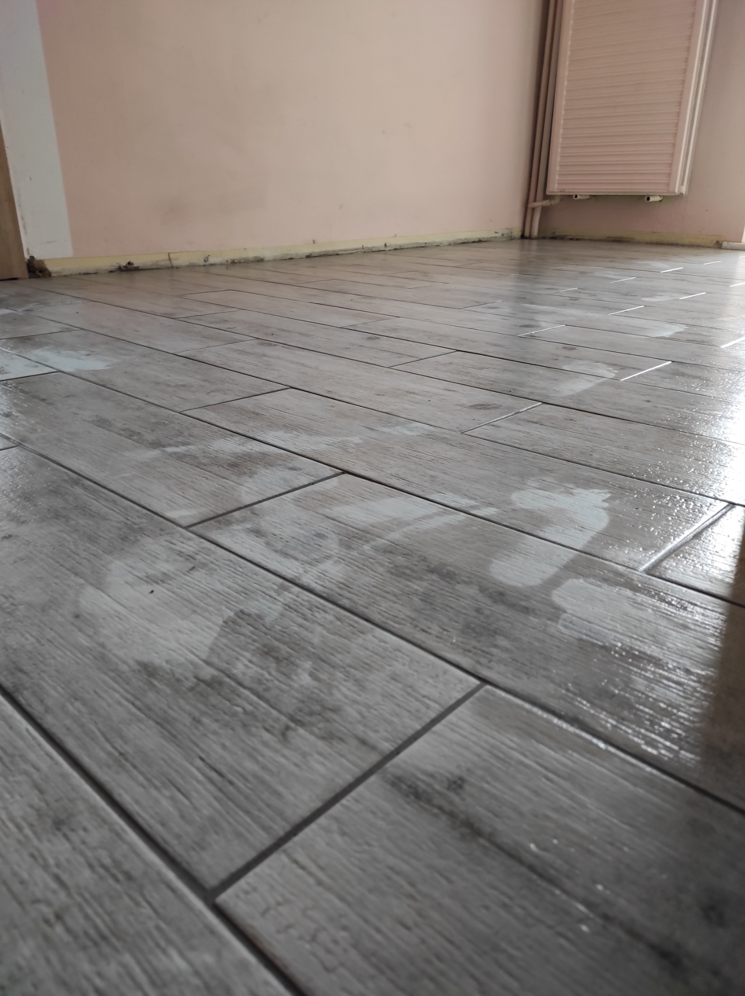 Ułożone szare płytki podłogowe imitujące drewno, świeżo po umyciu, z widocznymi smugami wody, na tle ściany w kolorze brudnego różu i grzejnika.