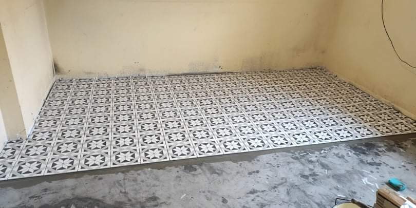 Fragment podłogi z ułożonymi płytkami ceramicznymi o wzorze geometrycznym w odcieniach szarości i bieli, widoczny świeży klej i narzędzia glazurnicze w rogu pomieszczenia.