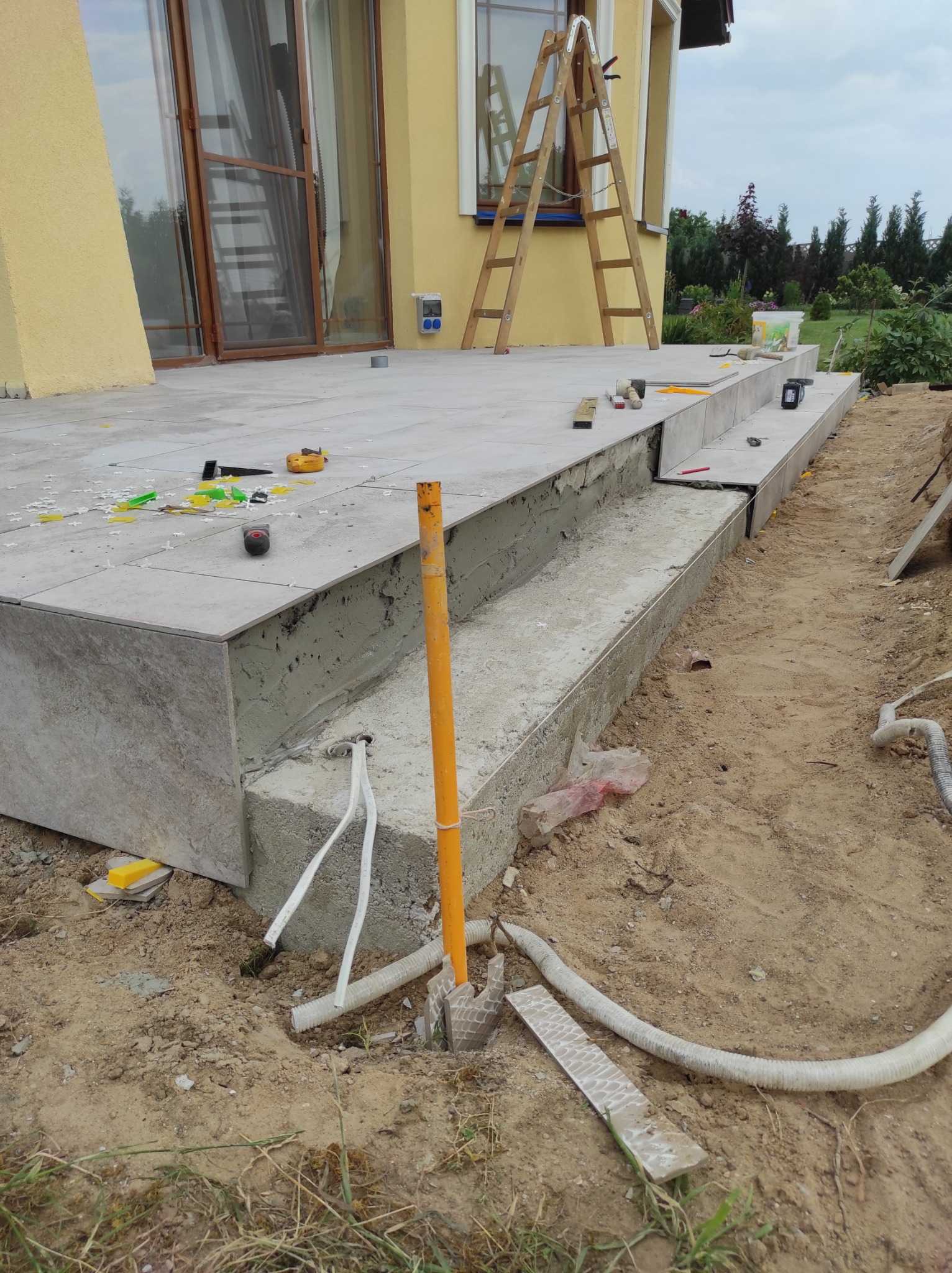 Układanie płytek gresowych na schodach zewnętrznych tarasu, widoczne narzędzia glazurnicze i elementy mocujące, drabina oparta o elewację budynku.