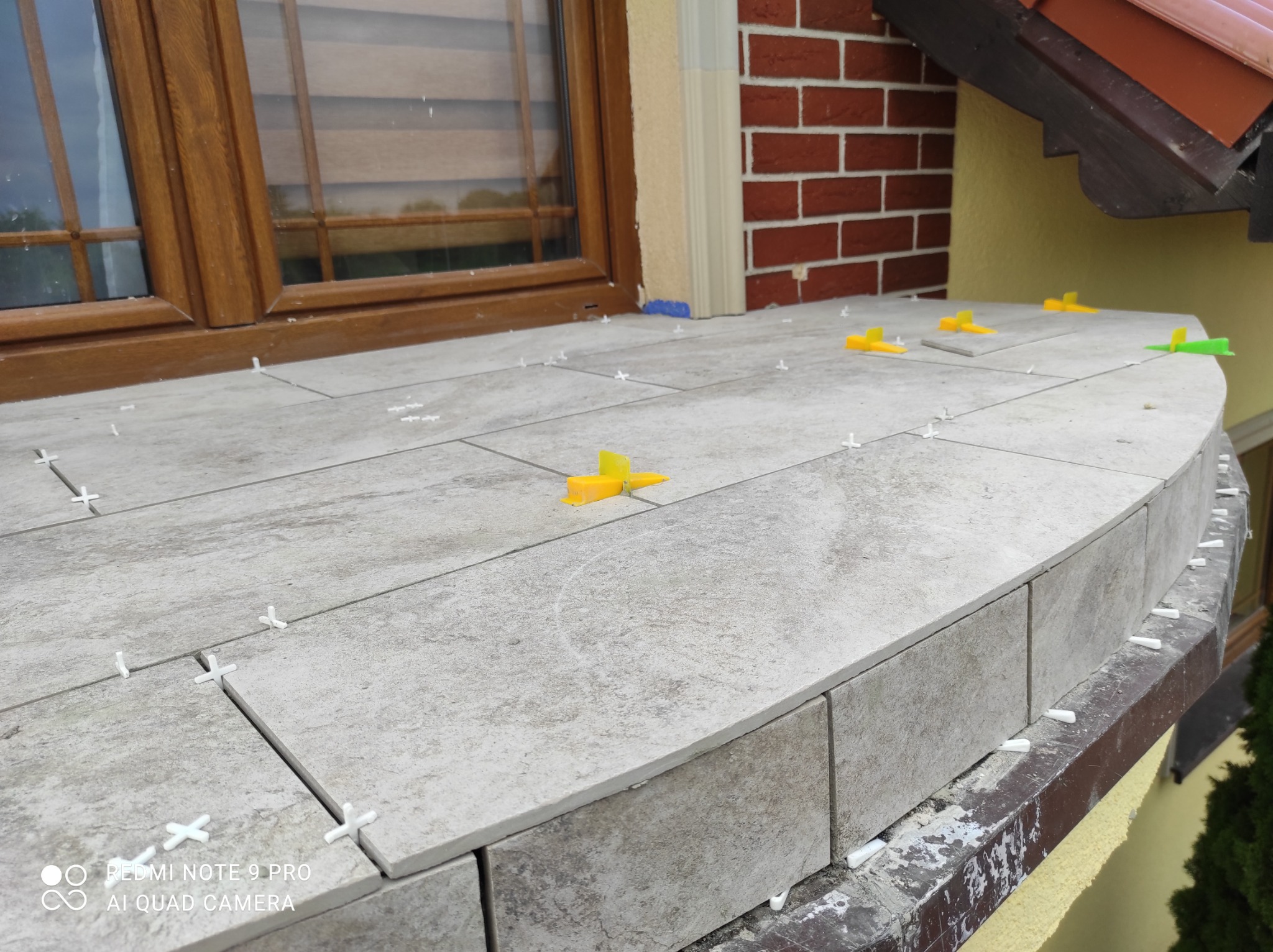 Kładzenie płytek na zaokrąglonym balkonie z użyciem krzyżyków dystansowych i klinów poziomujących, widoczne okno z drewnianą ramą i ceglany fragment elewacji.