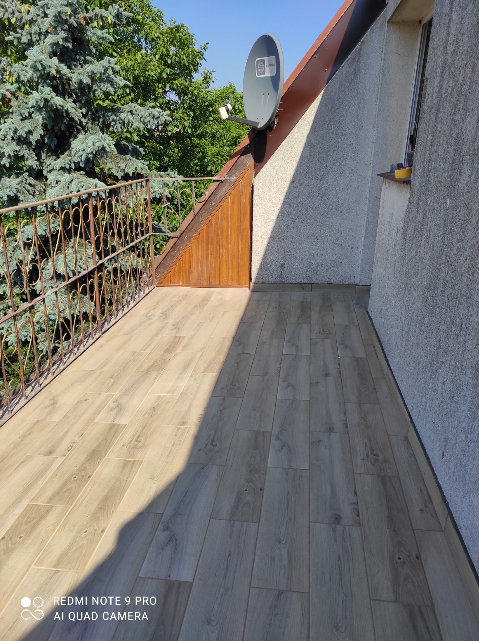 Balkon z nową podłogą z płytek imitujących drewno, żeliwna balustrada z ozdobnym wzorem, widok na drzewa i antenę satelitarną.