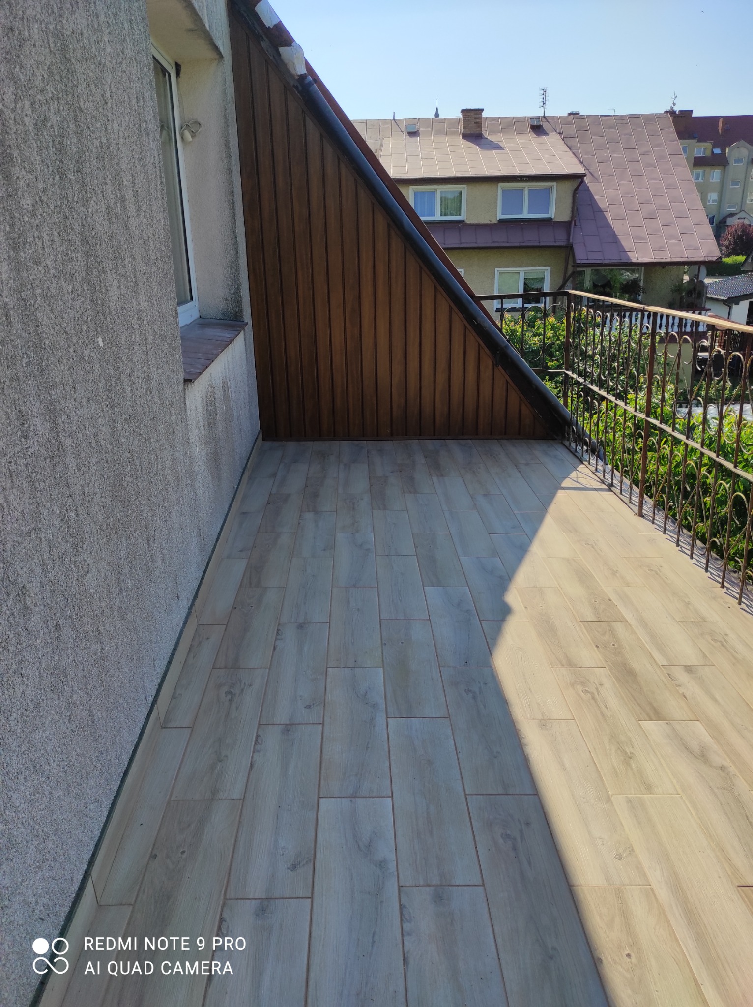 Balkon po remoncie z drewnopodobną podłogą z płytek, brązową boazerią na ścianie szczytowej i ozdobną metalową balustradą.