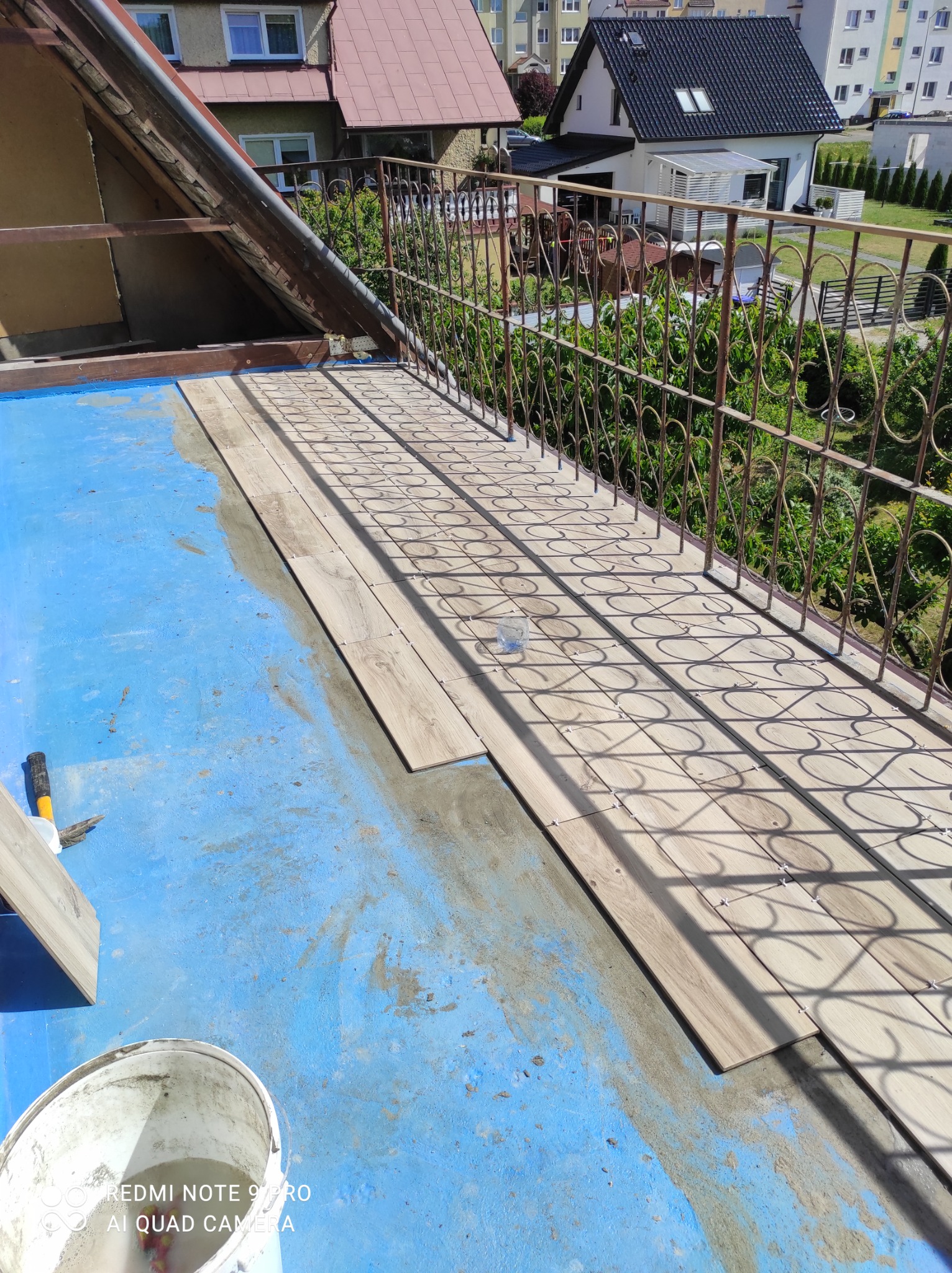 Balkon w trakcie układania płytek imitujących drewno na niebieskiej hydroizolacji, widoczne krzyżyki dystansowe i narzędzia, w tle balustrada i zabudowa domów.