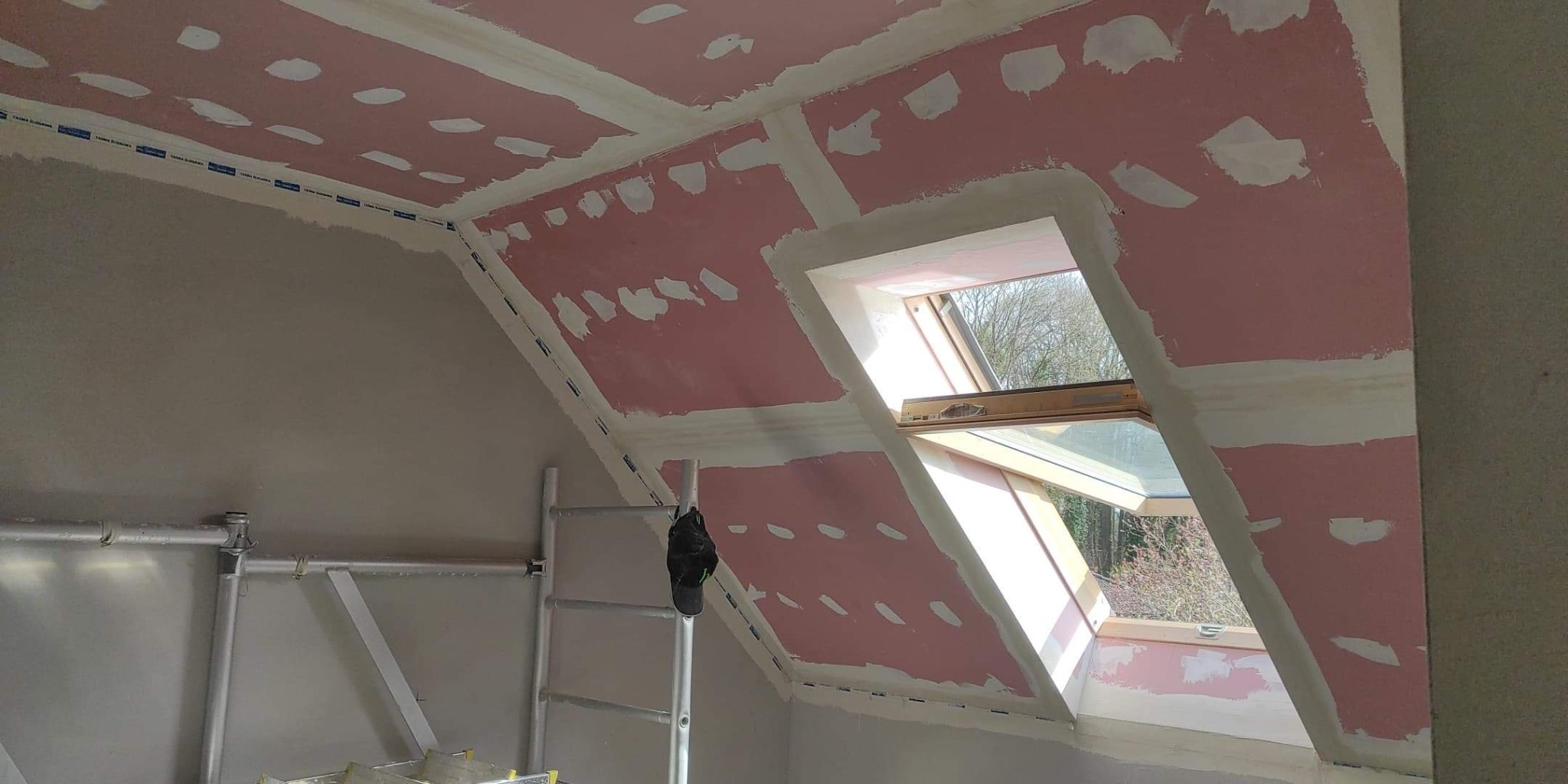 Poddasze w trakcie ocieplania, widoczne płyty gipsowo-kartonowe z zaszpachlowanymi łączeniami i otwartym oknem dachowym, drabina malarska w rogu pomieszczenia.
