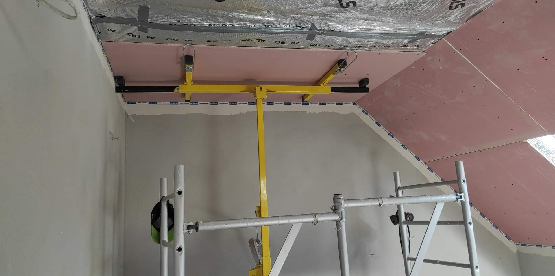 Montaż płyt gipsowo-kartonowych na poddaszu z użyciem żółtego podnośnika, widoczne rusztowanie i izolacja termiczna.