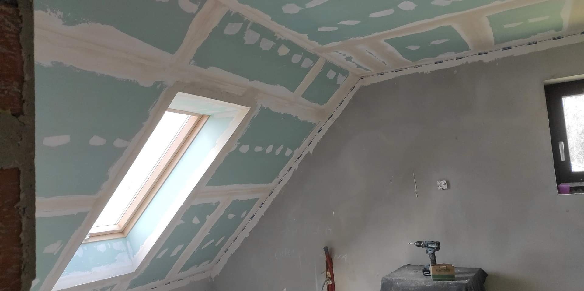 Poddasze w trakcie wykańczania: widoczne płyty gipsowo-kartonowe na skosach sufitu i ścian, szpachlowane łączenia, okno dachowe i narzędzia (wiertarka) na tle surowej ściany.
