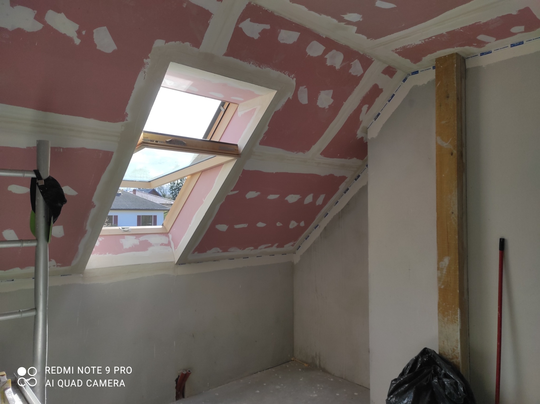 Poddasze w trakcie prac wykończeniowych: widoczne płyty gipsowo-kartonowe na skosach dachu, częściowo zaszpachlowane, okno dachowe w drewnianej ramie, fragment drabiny malarskiej z lewej strony.
