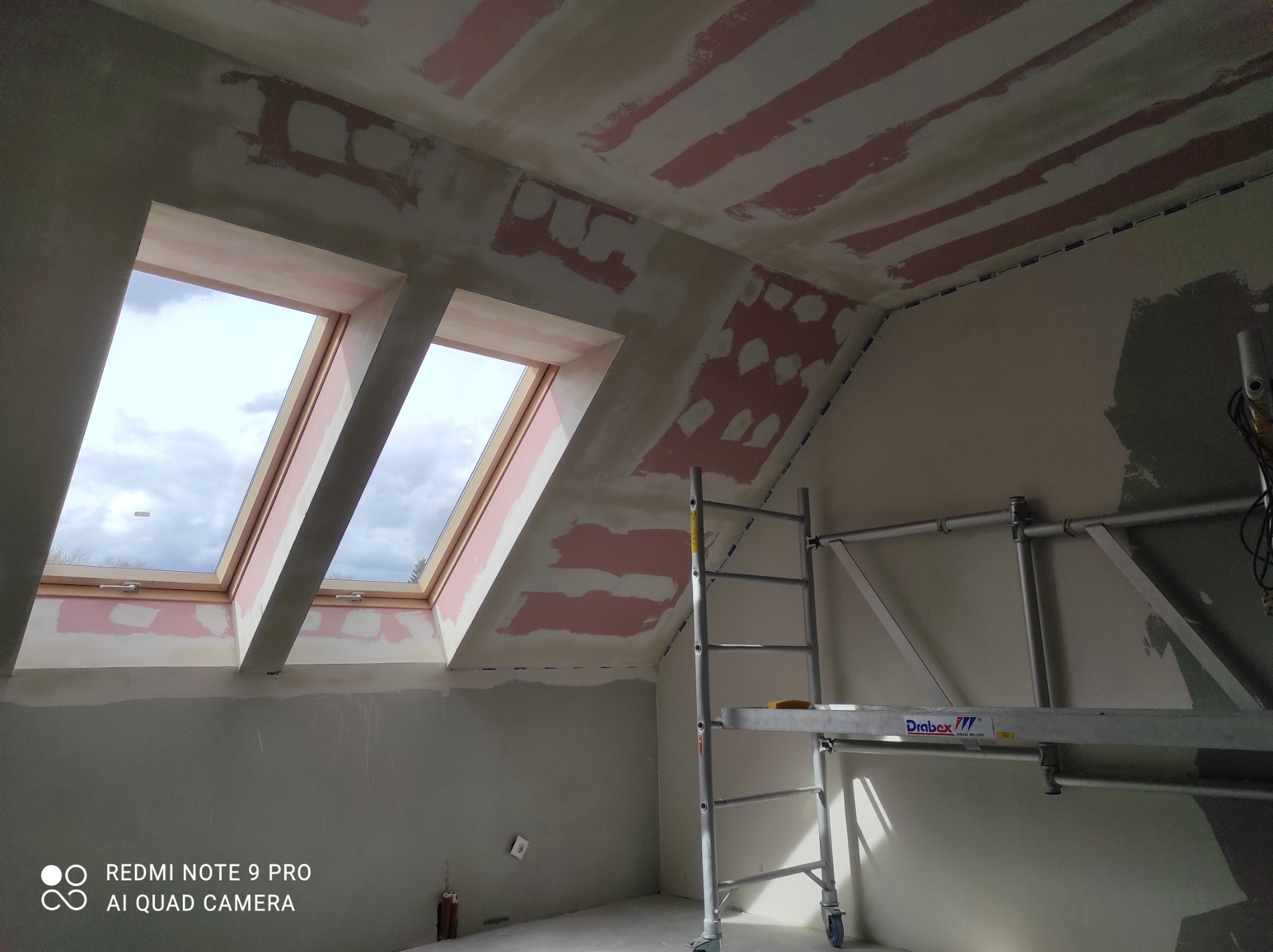 Poddasze w trakcie przygotowania do malowania, widoczne okna dachowe, ściany i sufit z fragmentami szpachli, rusztowanie drabinkowe.