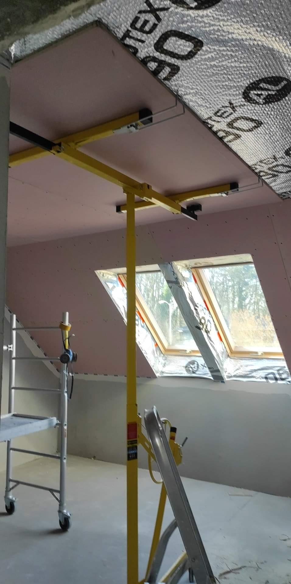 Montaż płyty kartonowo-gipsowej na poddaszu z oknami dachowymi, widoczny podnośnik do płyt i rusztowanie, folia termoizolacyjna na skosach dachu.