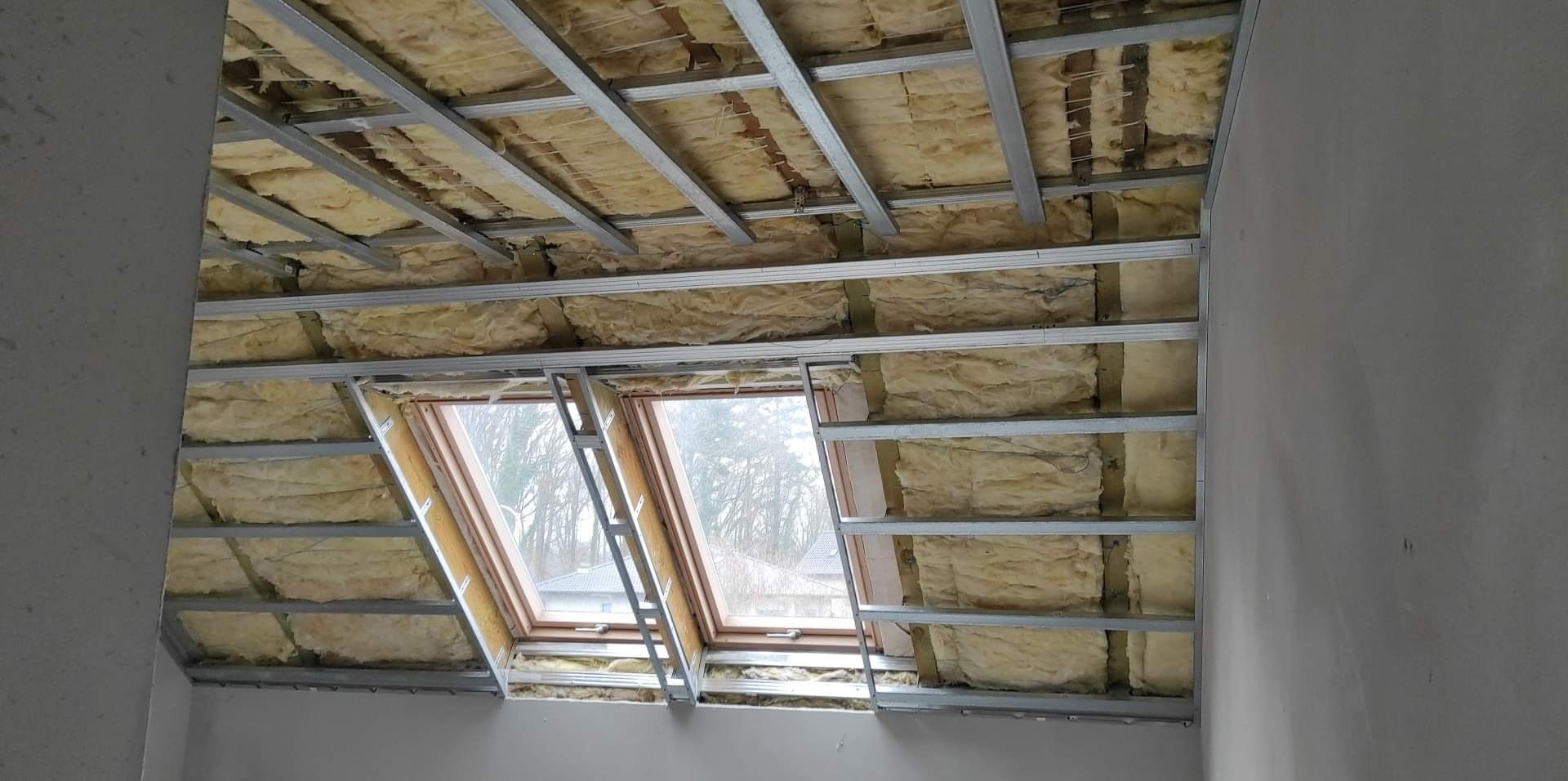 Poddasze w trakcie ocieplania z widoczną izolacją z wełny mineralnej, metalową konstrukcją pod płyty gipsowo-kartonowe i dwoma oknami dachowymi.