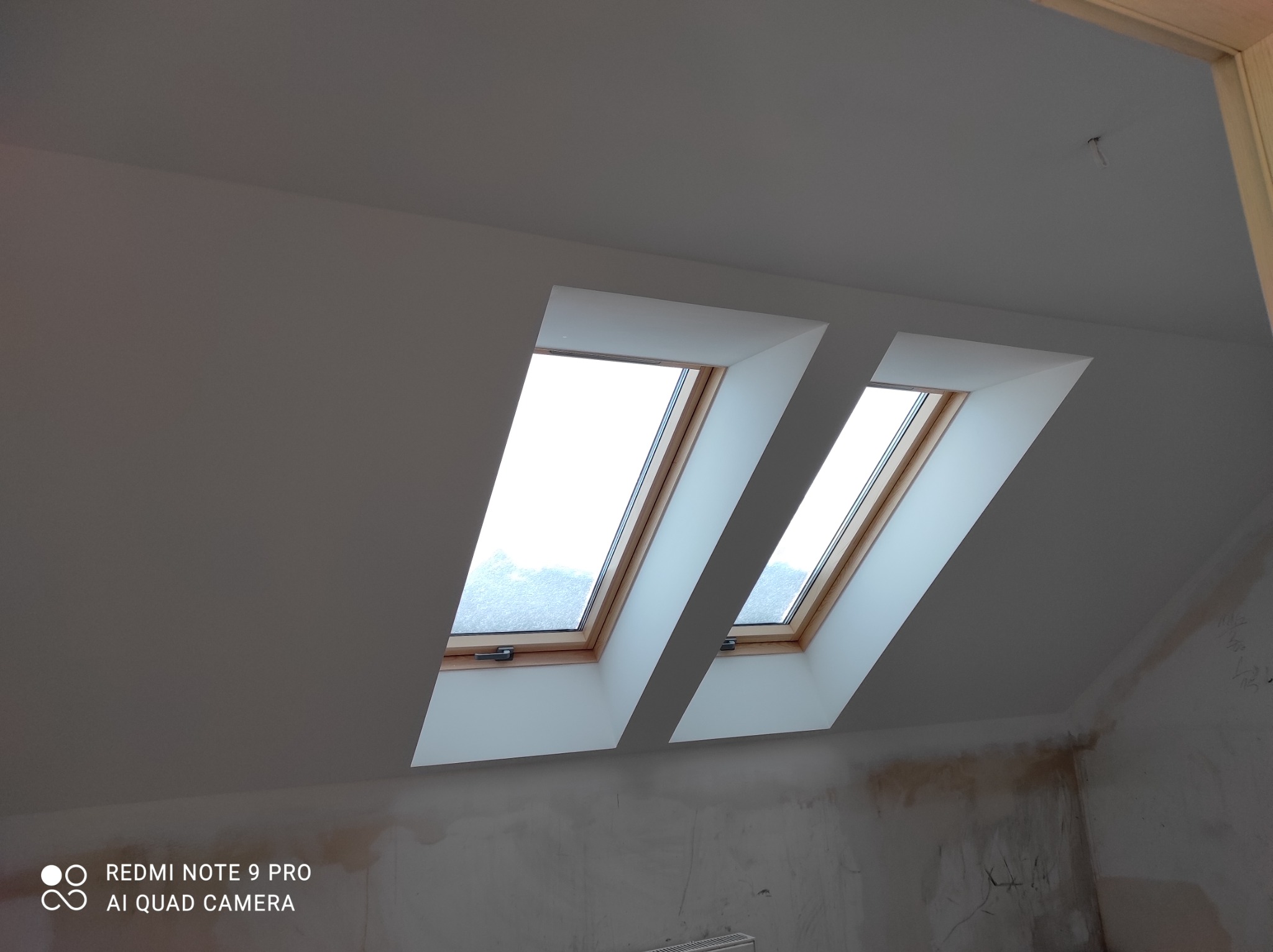 Dwa okna dachowe z drewnianą ramą zamontowane w białym, skośnym suficie poddasza. Ściany w trakcie malowania, widoczne ślady po szpachlowaniu.