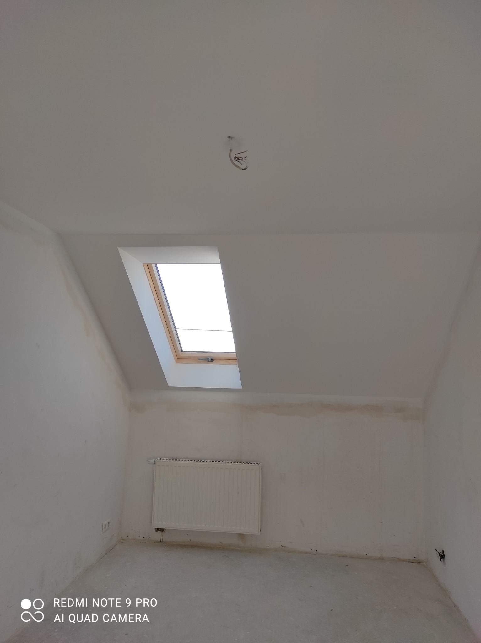 Poddasze w trakcie remontu, widoczne okno dachowe, grzejnik i ślady po naprawach na ścianach.