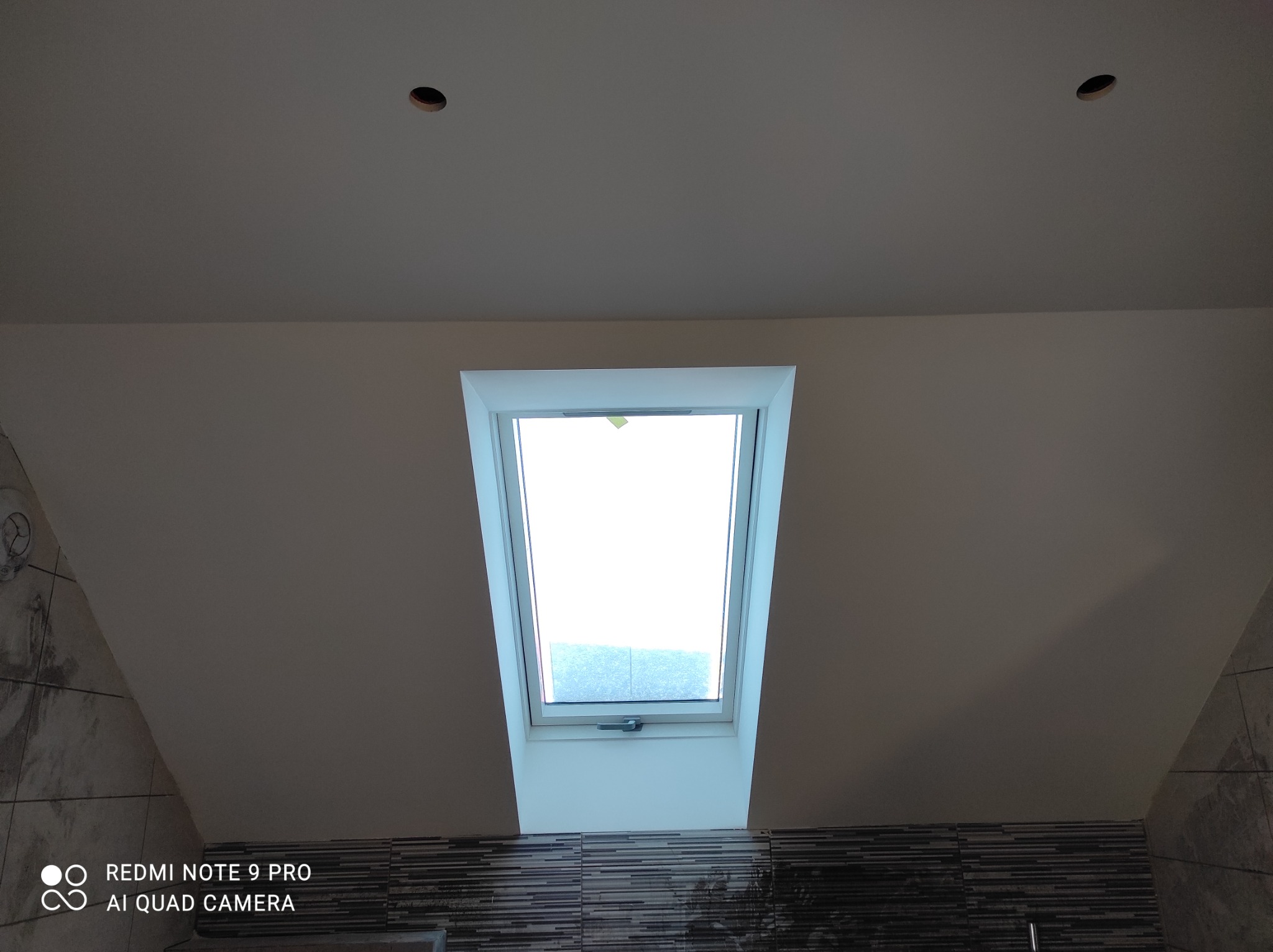 Poddasze łazienki z oknem dachowym, widoczne otwory na oświetlenie w suficie i dekoracyjne płytki na ścianie.