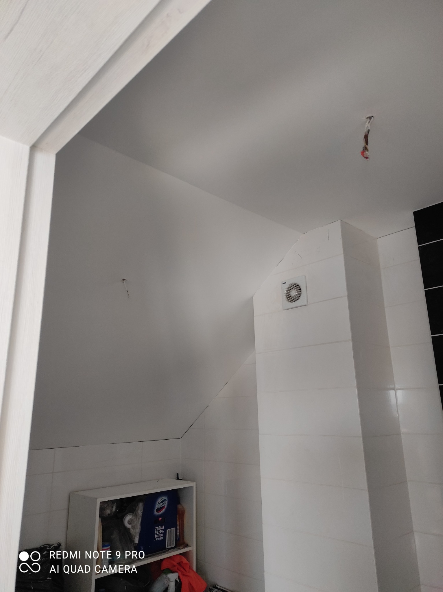 Łazienka po remoncie: białe płytki na ścianach i zabudowie, wentylator w narożniku, pod skosem sufitu półka z artykułami chemicznymi, wystające kable z sufitu.