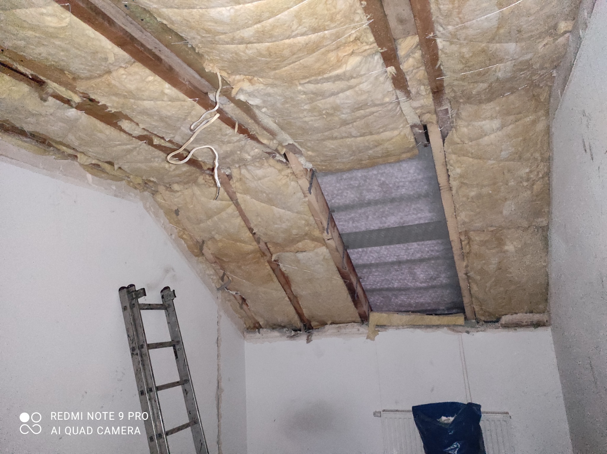 Poddasze w trakcie ocieplania wełną mineralną, widoczne drewniane belki konstrukcyjne, fragment schodów drabinkowych i odsłonięty fragment dachu z szarą blachą falistą.