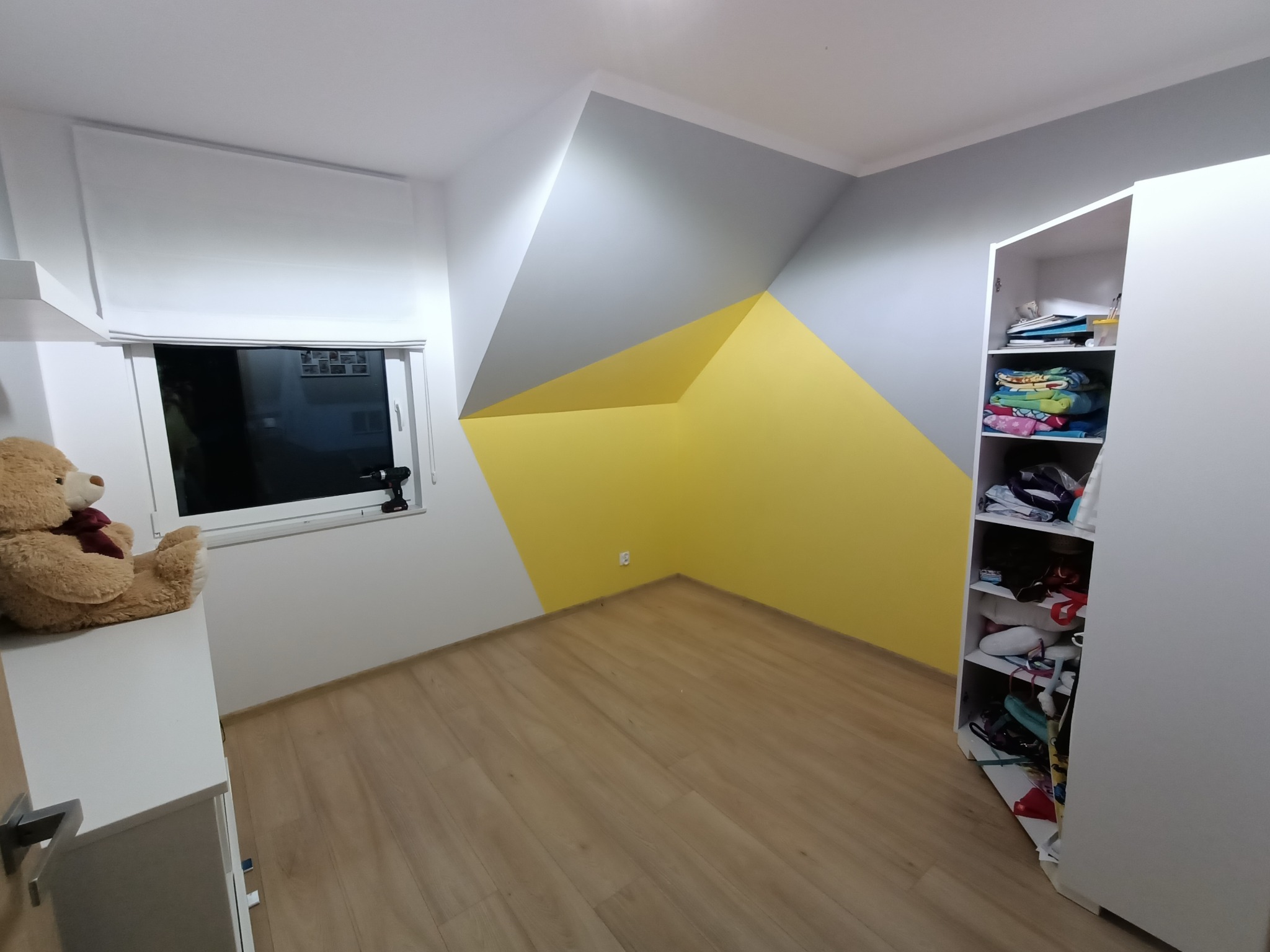 Pokój dziecięcy po remoncie: ściany pomalowane w geometryczne wzory w kolorach szarym i żółtym, jasne panele podłogowe, biała szafa i pluszowy miś na komodzie pod oknem.
