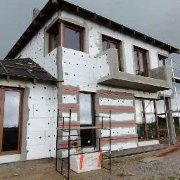 Ocieplanie elewacji domu jednorodzinnego z widocznymi fragmentami styropianu i rusztowaniami, przygotowane pod dalsze prace wykończeniowe elewacji, pochmurne niebo w tle.
