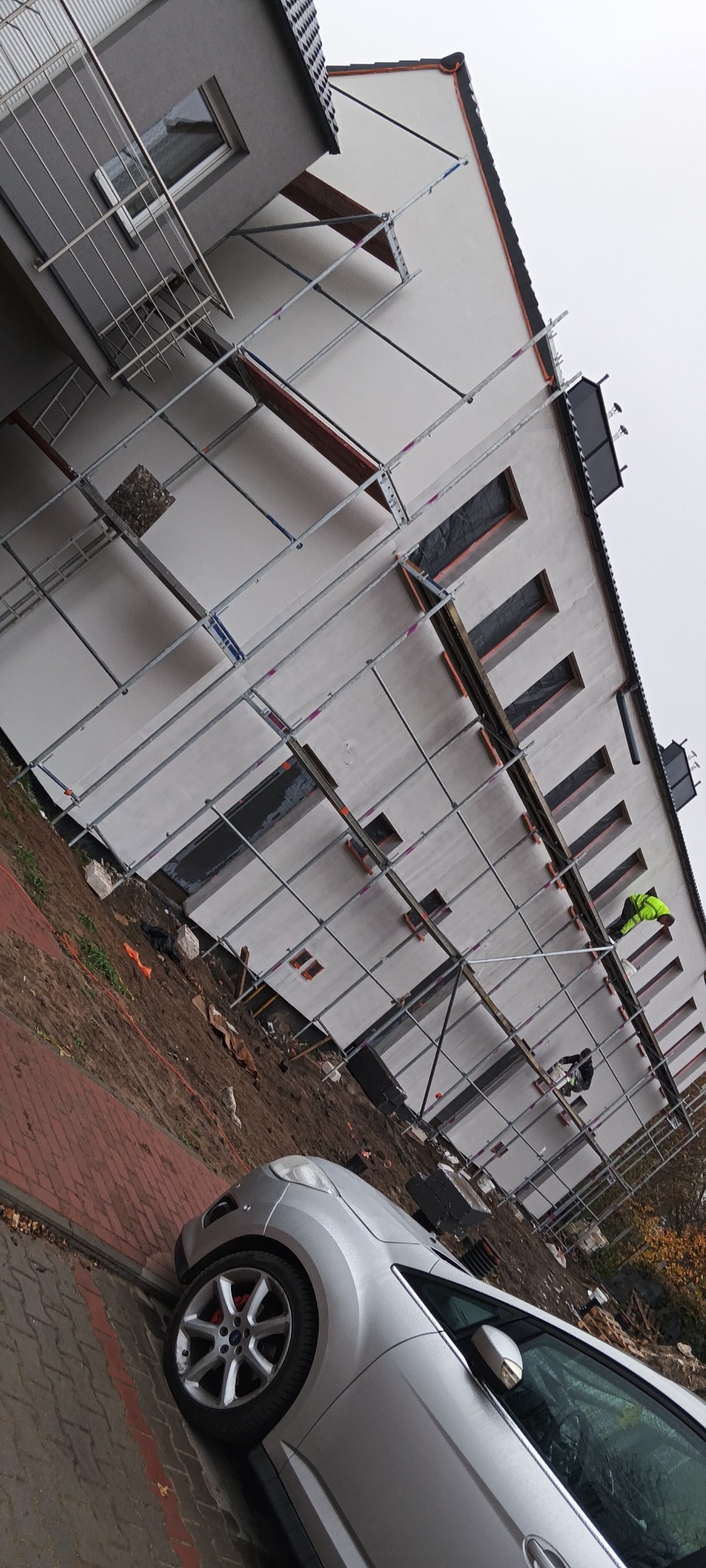 Budynek w trakcie prac elewacyjnych, widoczne rusztowania, zabezpieczone okna, pracownicy w odblaskowych kamizelkach, fragment srebrnego samochodu na pierwszym planie.