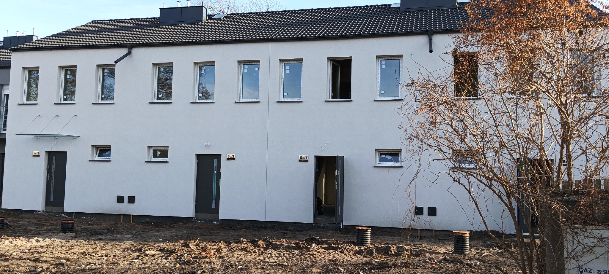 Nowoczesny budynek wielorodzinny z jasną elewacją, ciemną dachówką i czarnymi drzwiami wejściowymi, widoczne przygotowania do zagospodarowania terenu wokół budynku.