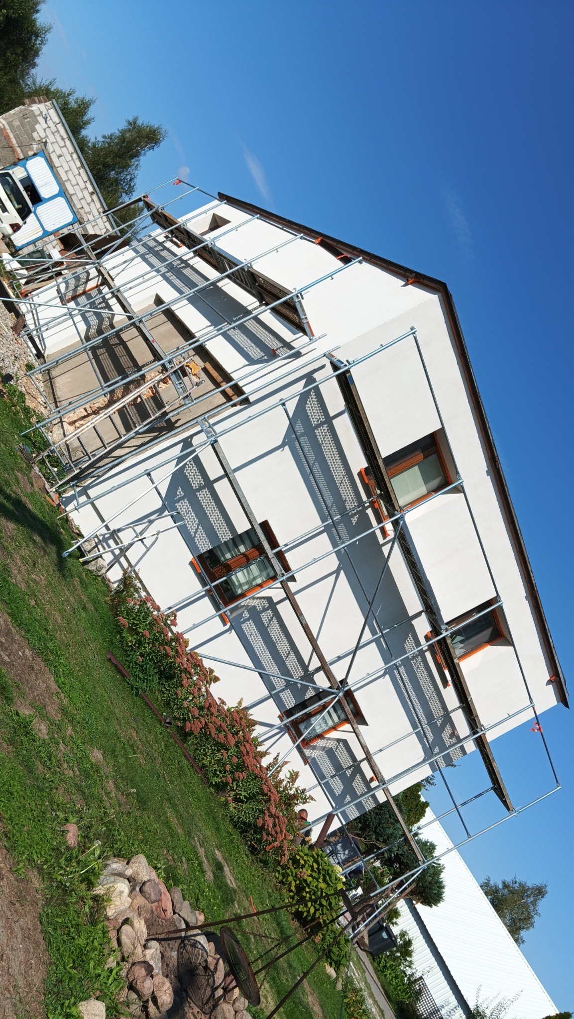 Dom jednorodzinny w trakcie prac elewacyjnych, widoczne rusztowania i świeżo pomalowana biała fasada, na pierwszym planie ogródek z kamieniami.