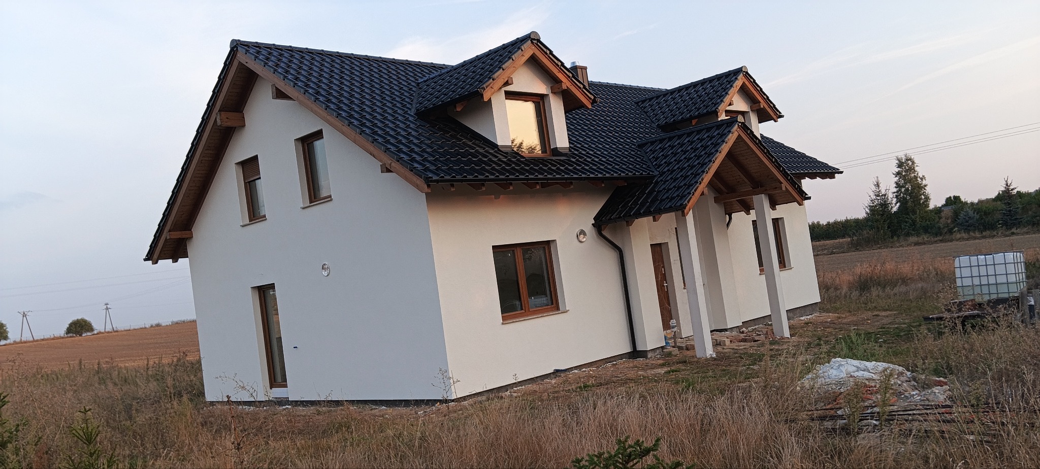 Nowoczesny dom jednorodzinny z białą elewacją, czarnym dachem i drewnianymi ramami okiennymi, usytuowany na skraju pola, z widocznym zbiornikiem na wodę w tle.