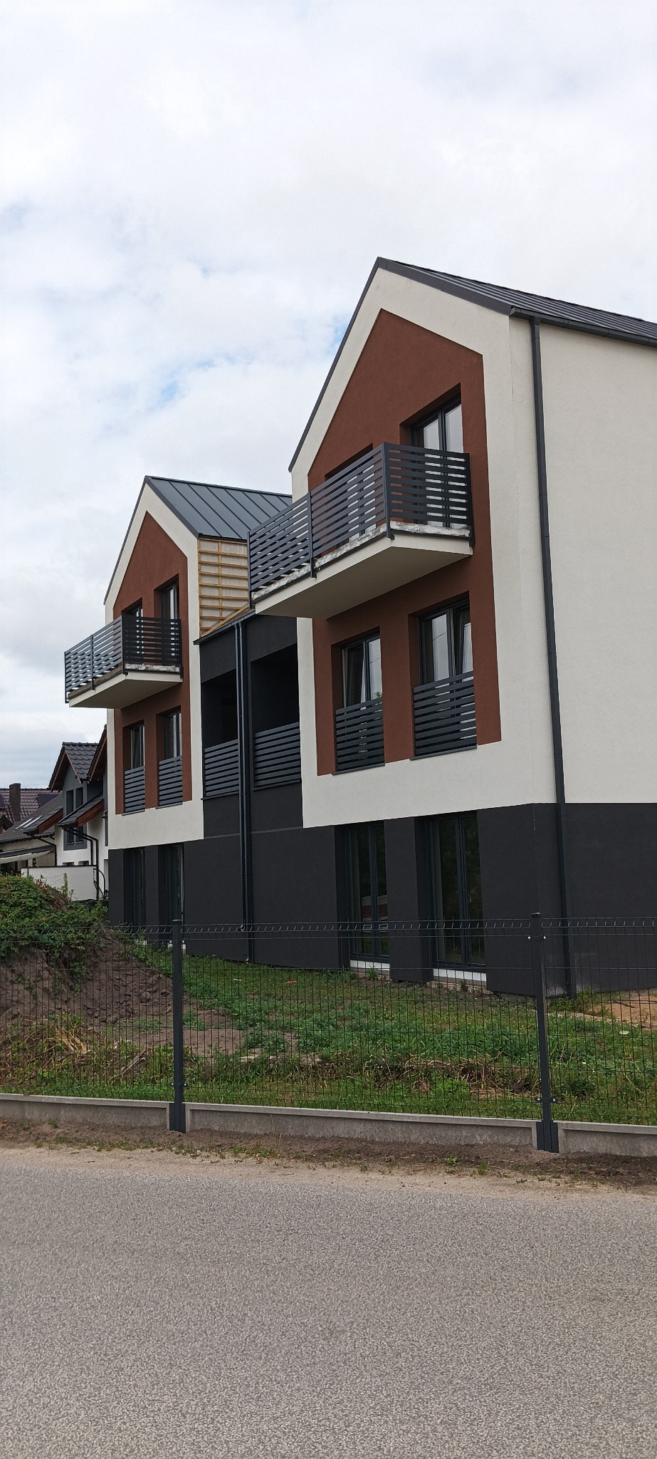 Nowoczesny budynek wielorodzinny z balkonami, elewacją w kolorach brązu, bieli i grafitu, widok z poziomu ulicy przez ogrodzenie.