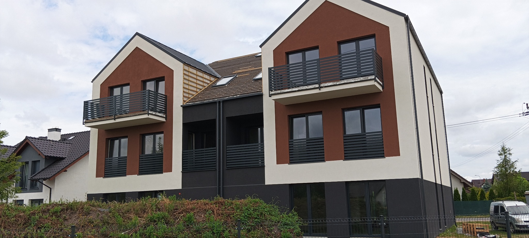 Nowoczesny budynek wielorodzinny z brązowo-białą elewacją, czarnymi oknami i balkonami, widoczne fragmenty dachu i sąsiednich budynków, pochmurne niebo.