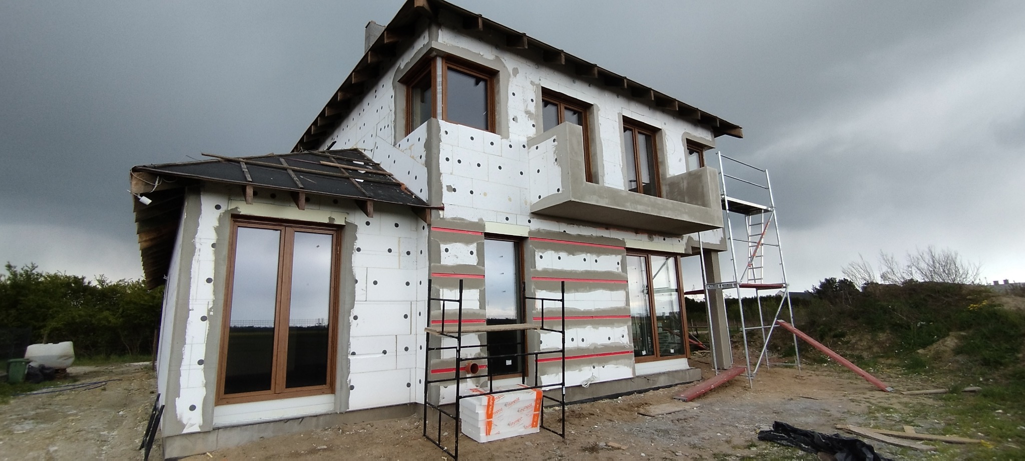 Ocieplanie elewacji domu jednorodzinnego z widocznymi fragmentami styropianu i rusztowaniami, przygotowane pod dalsze prace wykończeniowe elewacji, pochmurne niebo w tle.