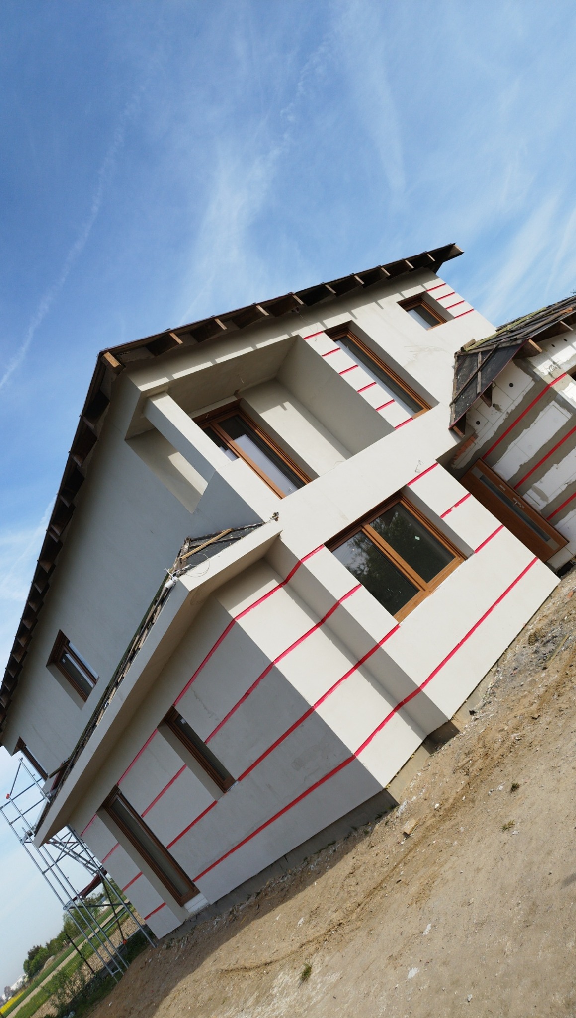 Dom w trakcie prac elewacyjnych, widoczne fragmenty ocieplenia oraz świeżo nałożony tynk z poziomymi liniami wyznaczającymi podział, rusztowanie z boku budynku, błękitne niebo w tle.