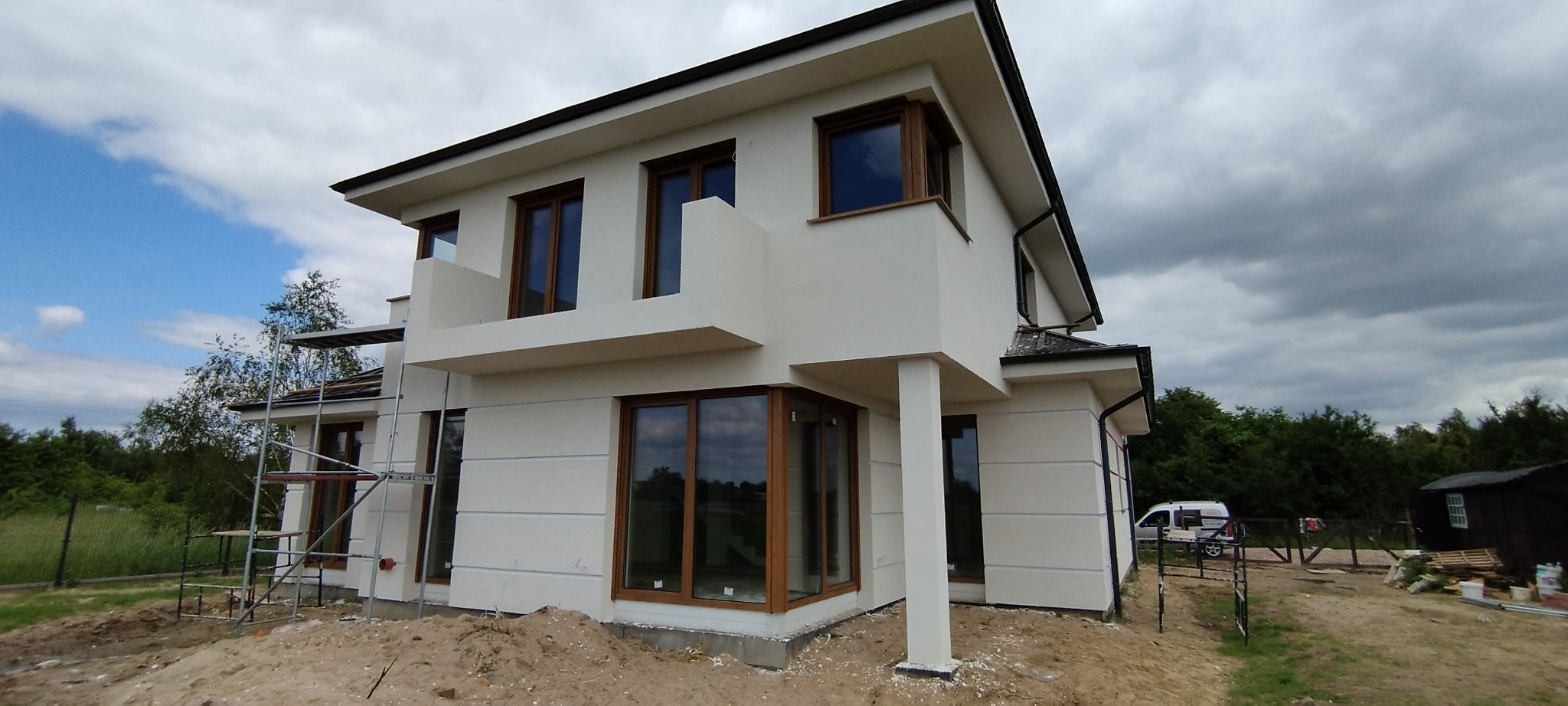 Nowoczesny dom jednorodzinny z jasną elewacją, drewnianymi oknami i balkonem, widoczny z zewnątrz na tle pochmurnego nieba, z fragmentem rusztowania po lewej stronie i samochodem dostawczym w tle.