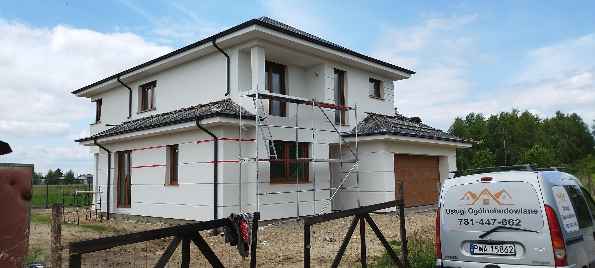 Dwukondygnacyjny dom jednorodzinny z białymi elewacjami w trakcie prac wykończeniowych, widoczne rusztowania i taśmy malarskie, na drugim planie las i samochód dostawczy z reklamą usług...
