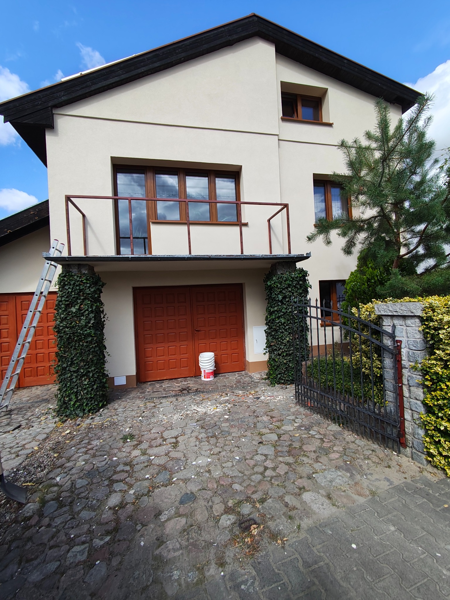 Jasna elewacja domu z balkonem, kolumny porośnięte bluszczem, drabina oparta o ścianę. Widoczny fragment bramy wjazdowej i kostka brukowa na podjeździe.