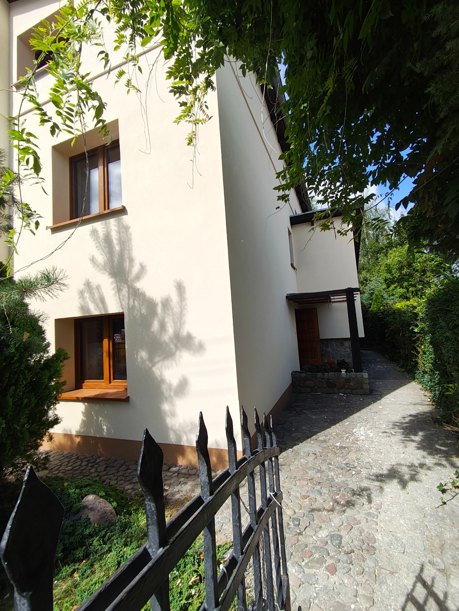 Elewacja domu jednorodzinnego w jasnym odcieniu, widoczna zza czarnego, kutego ogrodzenia. Na ścianie cień rzucany przez drzewo. Wejście do domu z kamienną ścieżką.