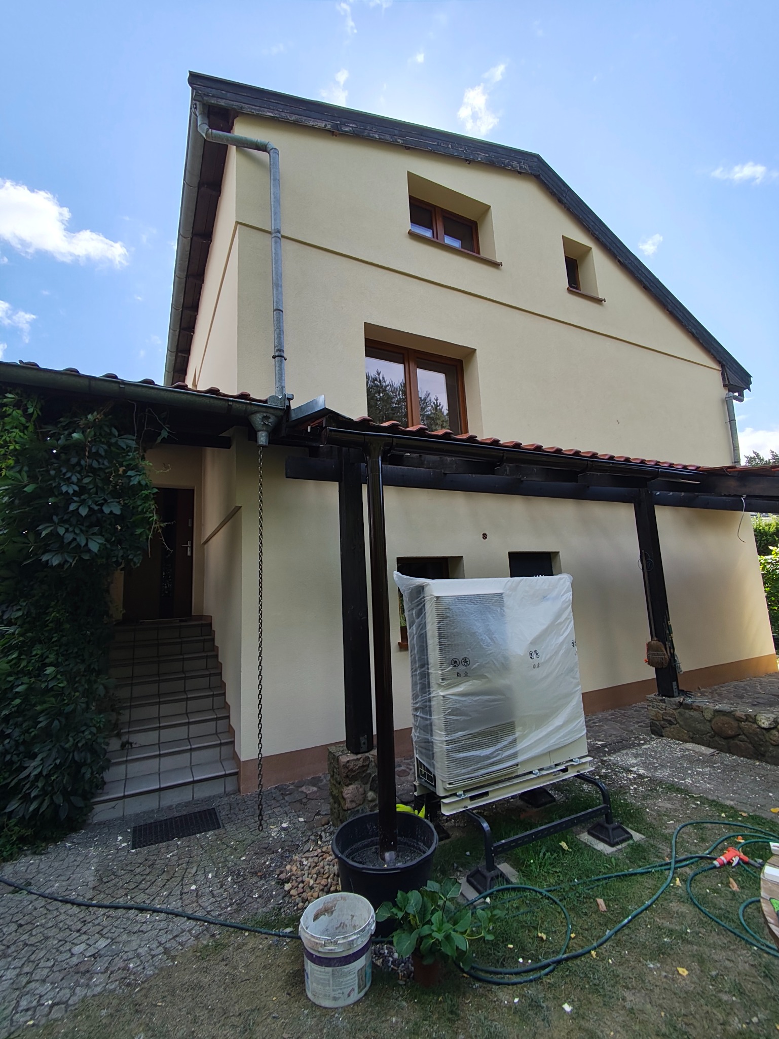 Dom z beżową elewacją, widoczne okna z brązowymi ramami. Z boku budynku stoi klimatyzator owinięty folią. Widoczny fragment ściany, schody i roślinność.
